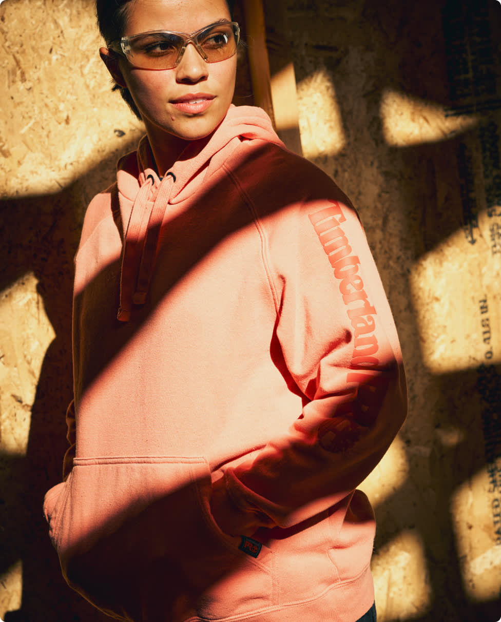 A women wearing a pink sweatshirt and safety glasses with a shaded plywood backdrop.