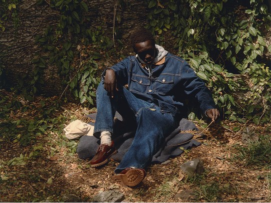 A man wearing all denim sitting outside on the ground in a wooded landscape.
