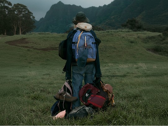 A person standing in a lush environment wearing and holding lots of backpacks. 