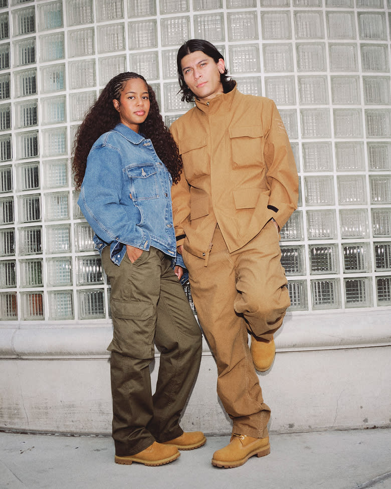 woman in denim jacket and green pants and brown boots standing next to man in brown clothing and brown boots