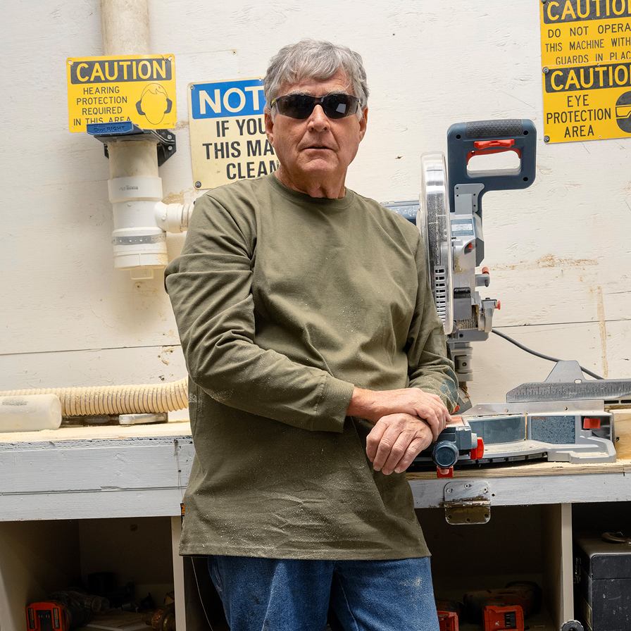 A man standing in a workshop with his arms crossed, wearing sunglasses and an olive green pullover sweatshirt. 