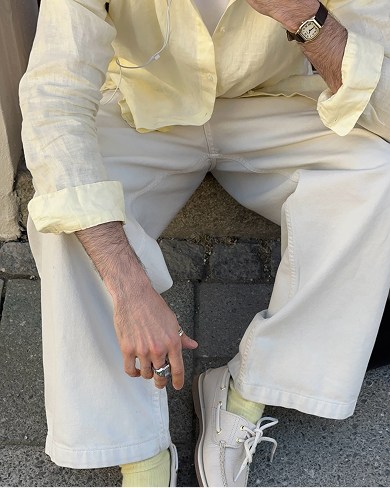 Close-up of off-white wide-leg trousers, light yellow socks, white leather boat shoes, and a hand with silver rings against a concrete pavement.