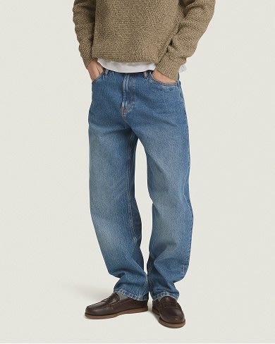 A person standing in wide-leg medium-wash jeans, a textured brown sweater, and dark brown loafers against a plain light background.