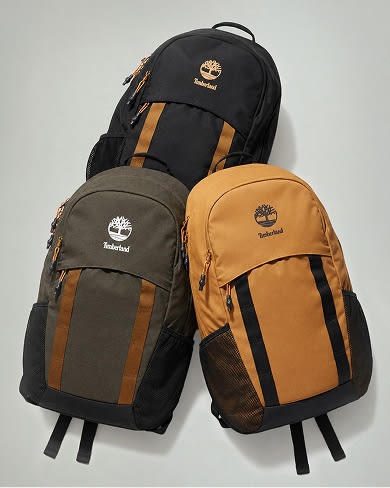 Three Timberland backpacks arranged against a neutral grey background: a black backpack at the top, an olive green backpack on the bottom left, and a tan backpack on the bottom right, each displaying the Timberland tree logo on the front upper compartment.