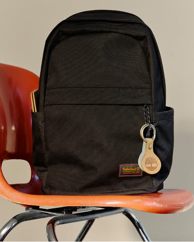 A black Timberland backpack resting on a vintage orange fiberglass chair, featuring a front zippered pocket with a small rectangular Timberland patch and a round beige leather key tag attached to the front ring.