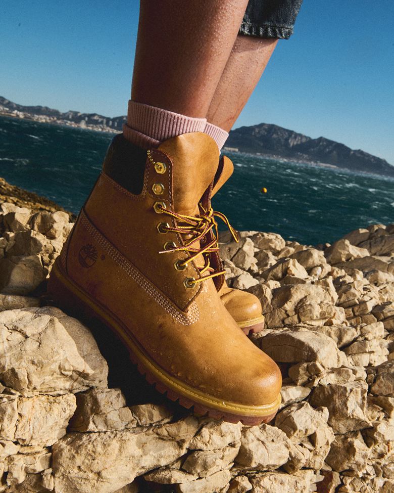 a closeup of a foot wearing the classic timberland boot standing on top of rocks at the beach