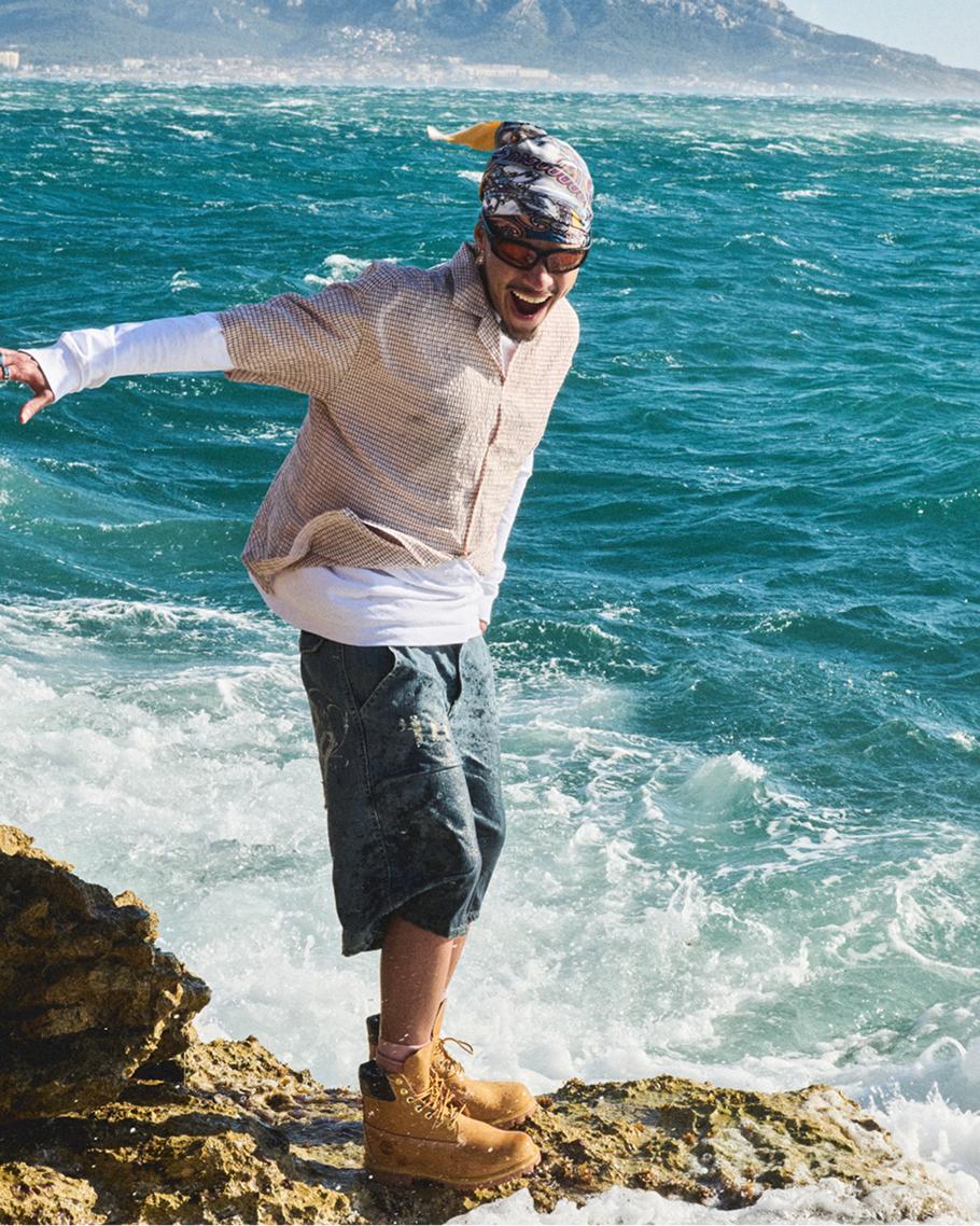 a man in summer clothing and timberland boots smiling at the beach standing on top of rocks