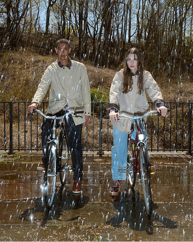 A man and a woman in light tan coats standing outdoors with their bicycles in the rain, with wet pavement reflecting their silhouettes and a wooded hill in the background.