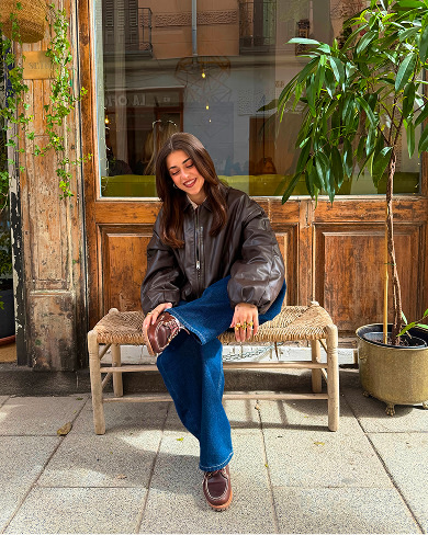 A smiling woman sitting outdoors on a woven bench in front of a wooden storefront, wearing a dark brown leather bomber jacket, wide-leg blue jeans, and brown leather boat shoes or lug-sole loafers.