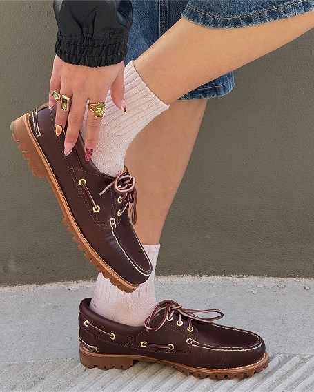 A close-up view of a person wearing dark brown leather 3-eye lug boat shoes with light-colored stitching and white ribbed socks, raising one foot and reaching down to touch the heel while wearing denim shorts and rings.