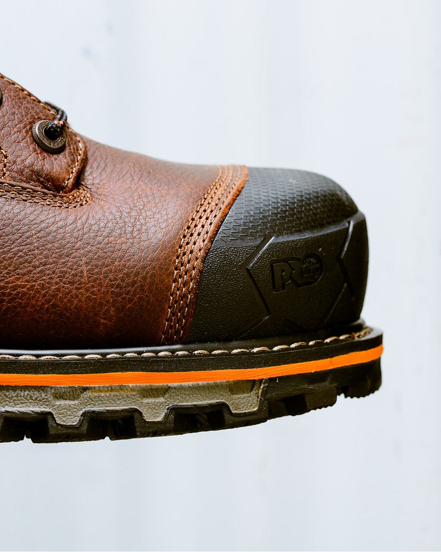 Extreme close-up profile view of the toe box on a brown leather Timberland PRO work boot, highlighting the dark rubber safety toe guard stamped with the PRO logo, double-stitched seams, and an orange accent line running along the thick rubber tread sole.