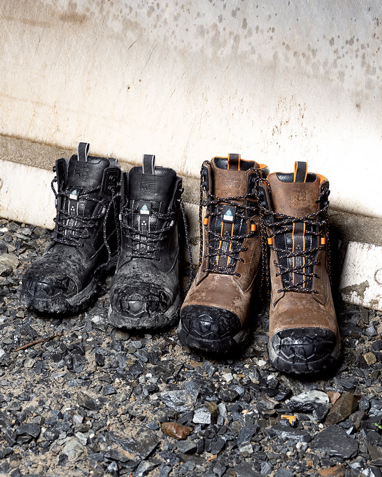 Two pairs of heavy-duty Timberland PRO work boots—one pair in black leather on the left and one pair in brown leather with orange accents on the right—standing upright on gray gravel against a concrete wall.