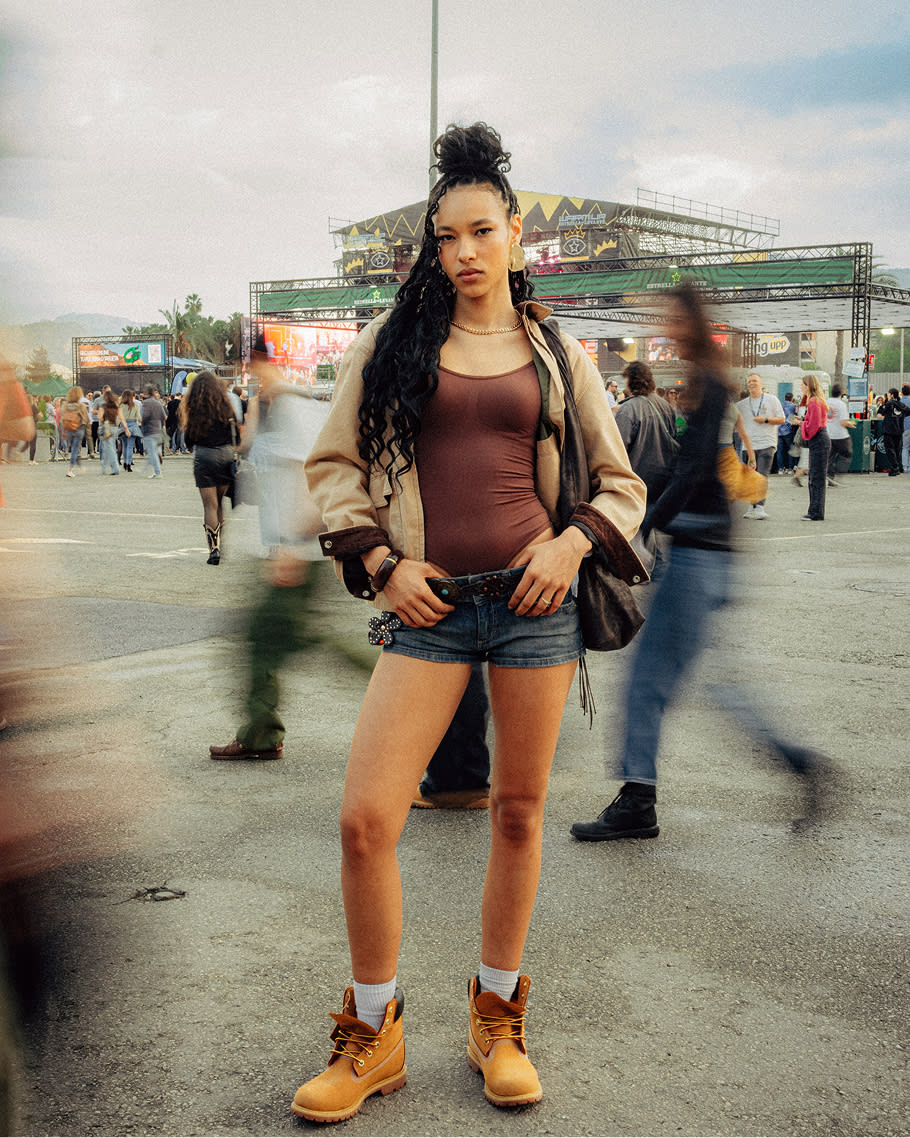 Full-length portrait of a stylish woman standing outdoors at an open-air event, styled in classic wheat Timberland boots, white socks, denim shorts, a brown bodysuit, and an unbuttoned tan jacket, with her hair in a high bun and long braids.