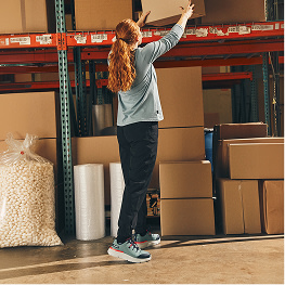 A women working wearing blue work sneakers stacking boxes in a warehouse setting.