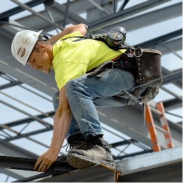 A man working on an I-Beam wearing a hard hat, highlighter yellow shirt, jeans and a pair of work boots.
