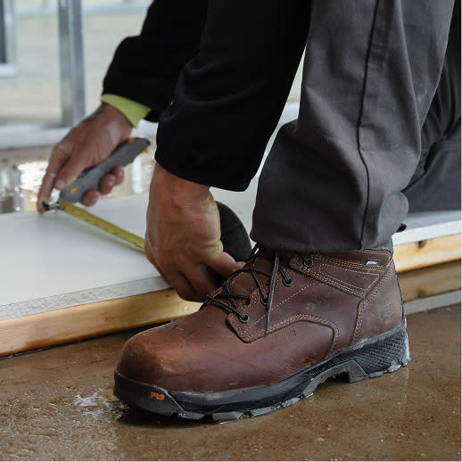 A person measuring a wooden board resting in a puddle. The persons leather brown work boot is in the foreground.