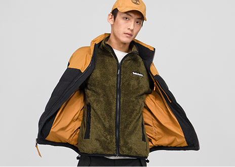 Image of a young man in an olive green vest, wheat colored Timberland tree logo baseball cap and a black and wheat colored jacket that he's holding open to show off the inside.