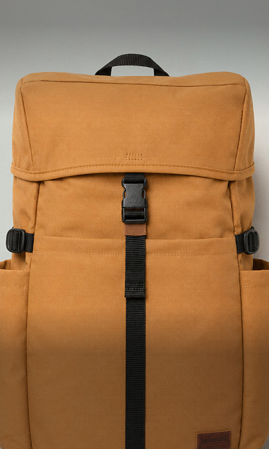 Front view of a tan canvas backpack featuring a top flap secured with a black plastic buckle, vertical black webbing strap down the front center, side compression straps, and side pockets.