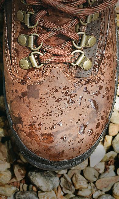 Top-down close-up view of a brown leather hiking boot covered in water droplets resting on a bed of light-colored pebbles, showing metal lace hooks and crisscrossed laces.