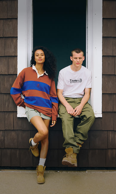 A woman and a man wearing Timberland footwear posing by a white-framed window on a dark brown shingled wall: the woman on the left stands leaning in a brown and blue striped rugby shirt, short skirt, white socks, and wheat Timberland boots; the man on the right sits on the windowsill wearing a white t-shirt, olive pants, and wheat Timberland boots.