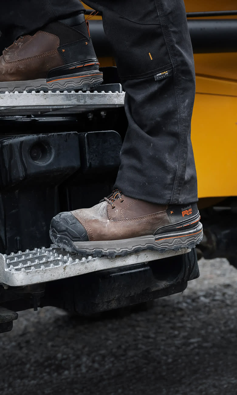 A close-up side view of a worker wearing dark trousers and brown Timberland PRO work boots stepping onto the metal grated steps of a yellow heavy construction vehicle.
