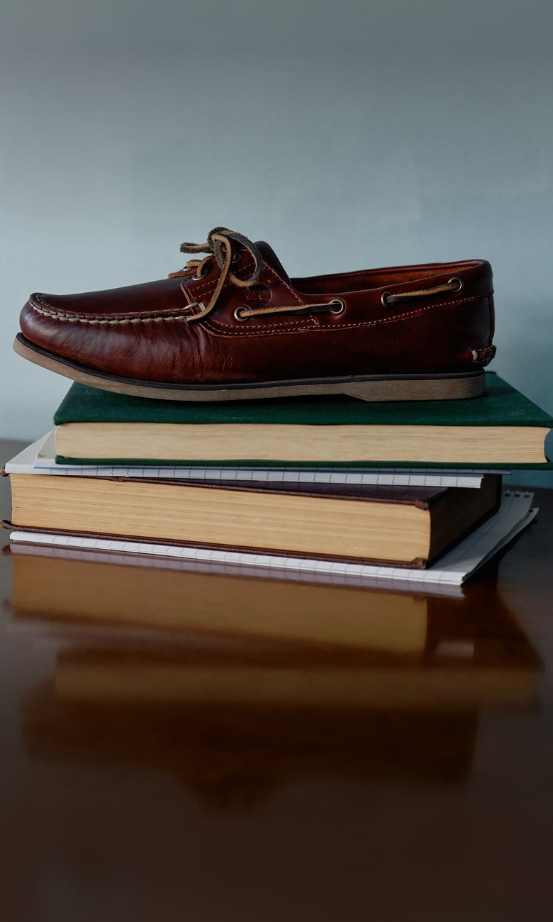 A single brown leather boat shoe resting on top of a stack of books and spiral notebooks placed on a reflective wooden table against a plain blue wall.