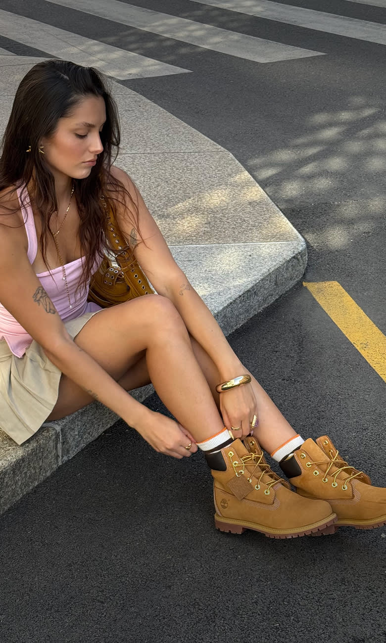 A young woman with long dark hair sits on a street curb near a crosswalk, adjusting her white crew socks worn with wheat-colored Timberland boots, paired with a light pink tank top and a beige mini skirt.