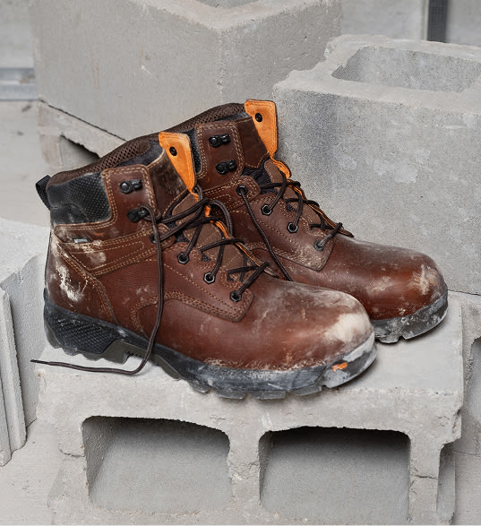 A pair of brown leather work boos, sitting upon a cinder block against a concrete backdrop. The boots have dried concrete splattered on them.